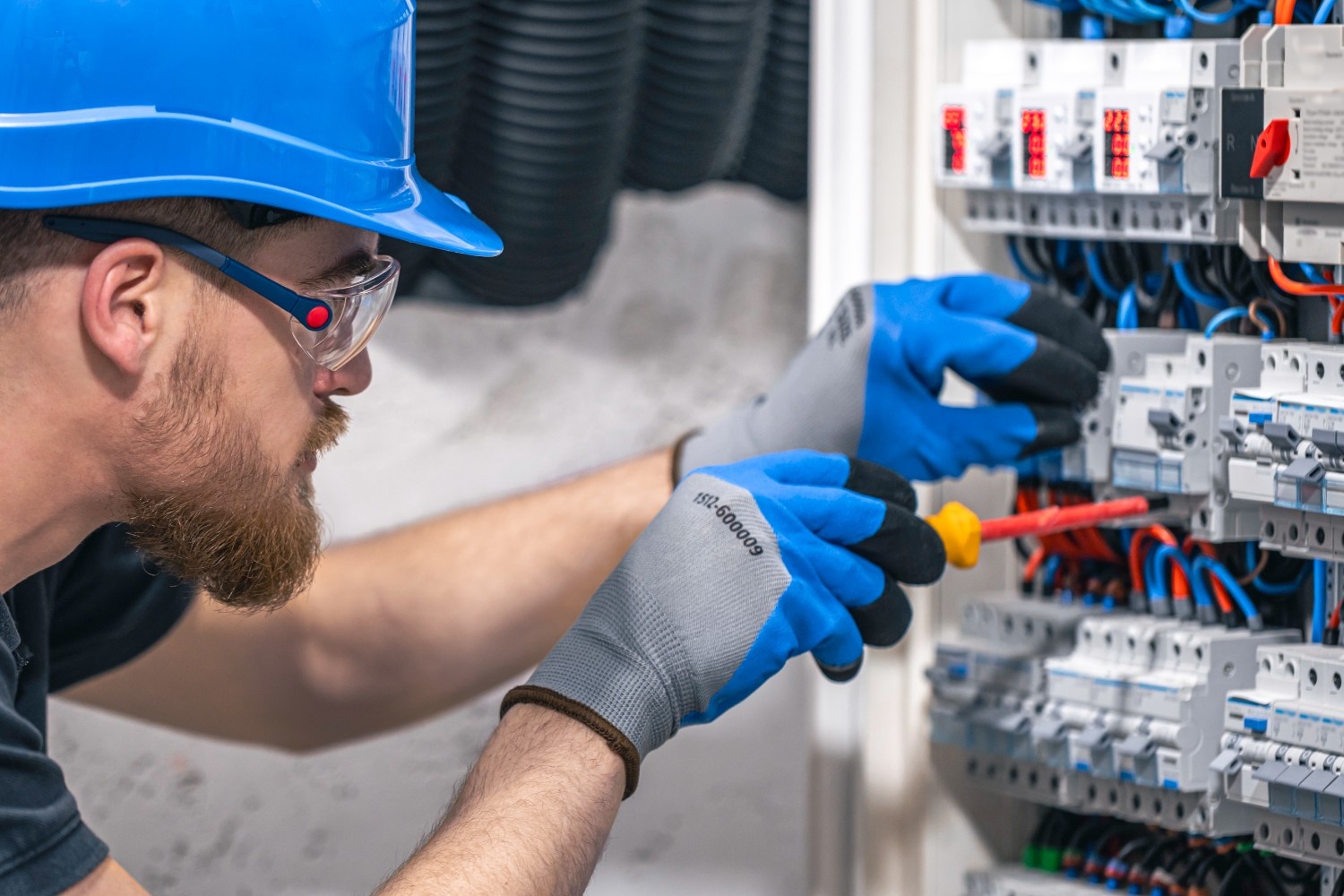 Electrician wearing high-grip work gloves using insulated screwdriver on electrical control panel – safety and precision gear in use.
